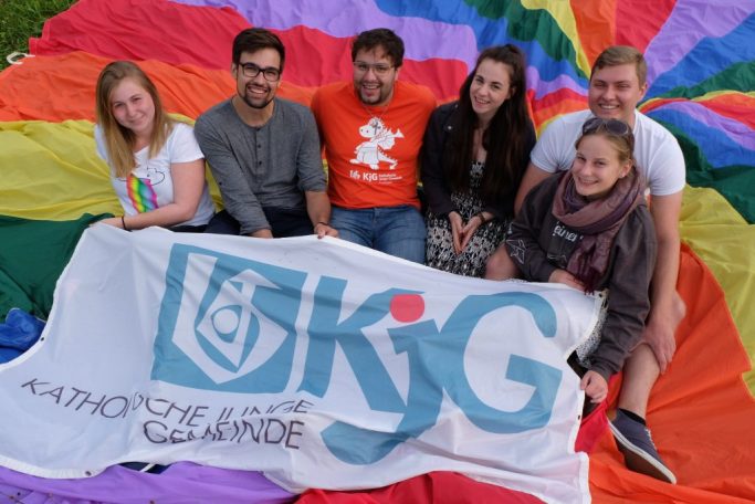 Sechs Personen posieren vor einem bunten Regenbogenbanner und einem KjG-Banner