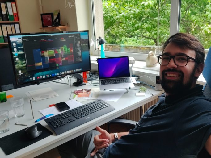 Felix sitzt lächeln an einem Schreibtisch mit zwei Bildschirmen und Pflanzen im Hintergrund.