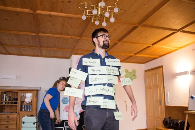 Mann mit Zetteln am Körper steht im Raum, während eine Frau im Hintergrund agiert.