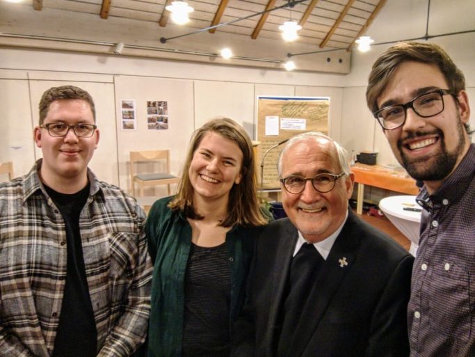 Drei Personen lächeln zusammen mit einem Bischof in einem Innenraum, mit Tischen und Stühlen im Hintergrund. 