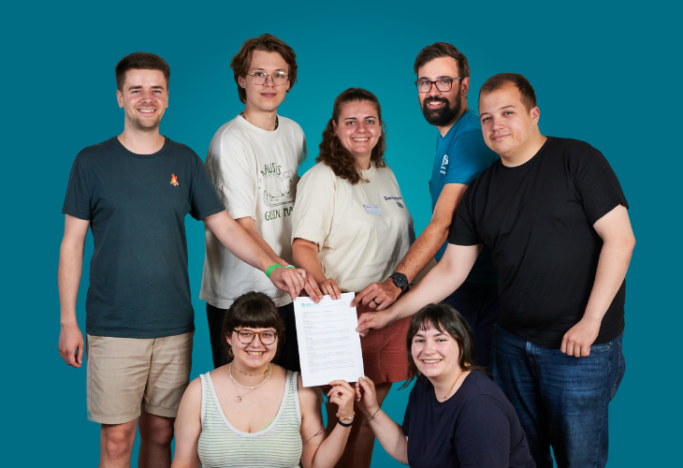 Gruppe von sieben Personen posiert gemeinsam und hält ein Dokument in der Mitte.