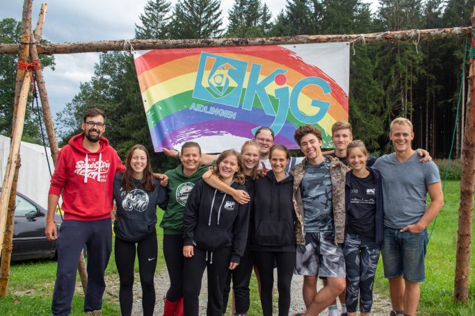 Gruppe von Jugendlichen vor einem bunten, regenbogenfarbenen Banner der KjG.
